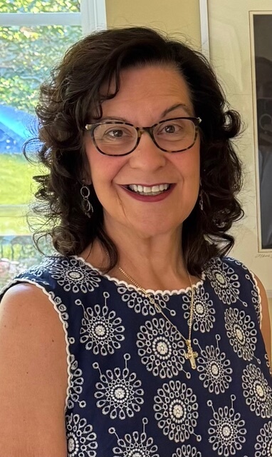Smiling woman with glasses in blue patterned dress, standing indoors with window light and framed artwork in background.