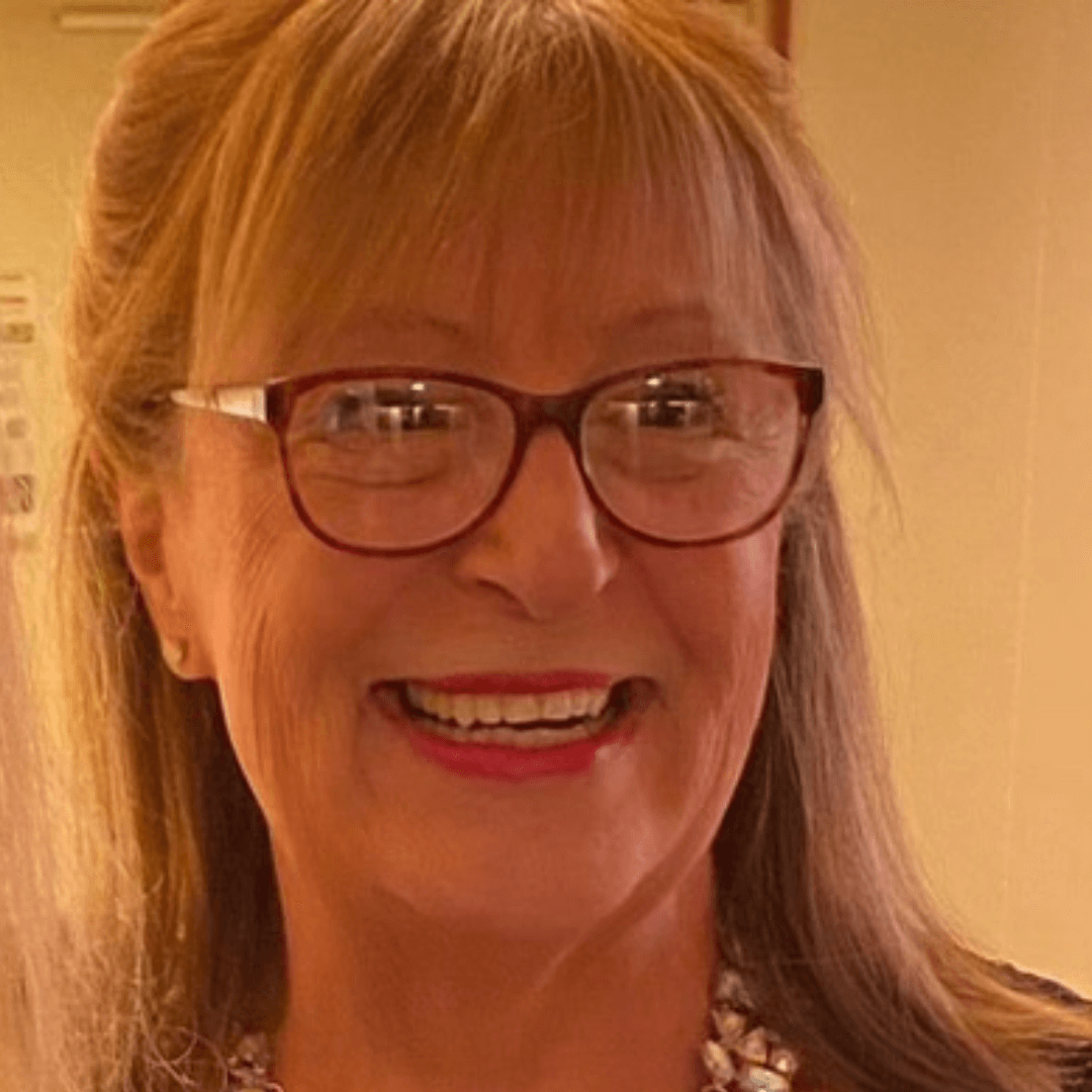 Smiling woman with glasses, long hair, and a necklace, indoors, warm lighting.