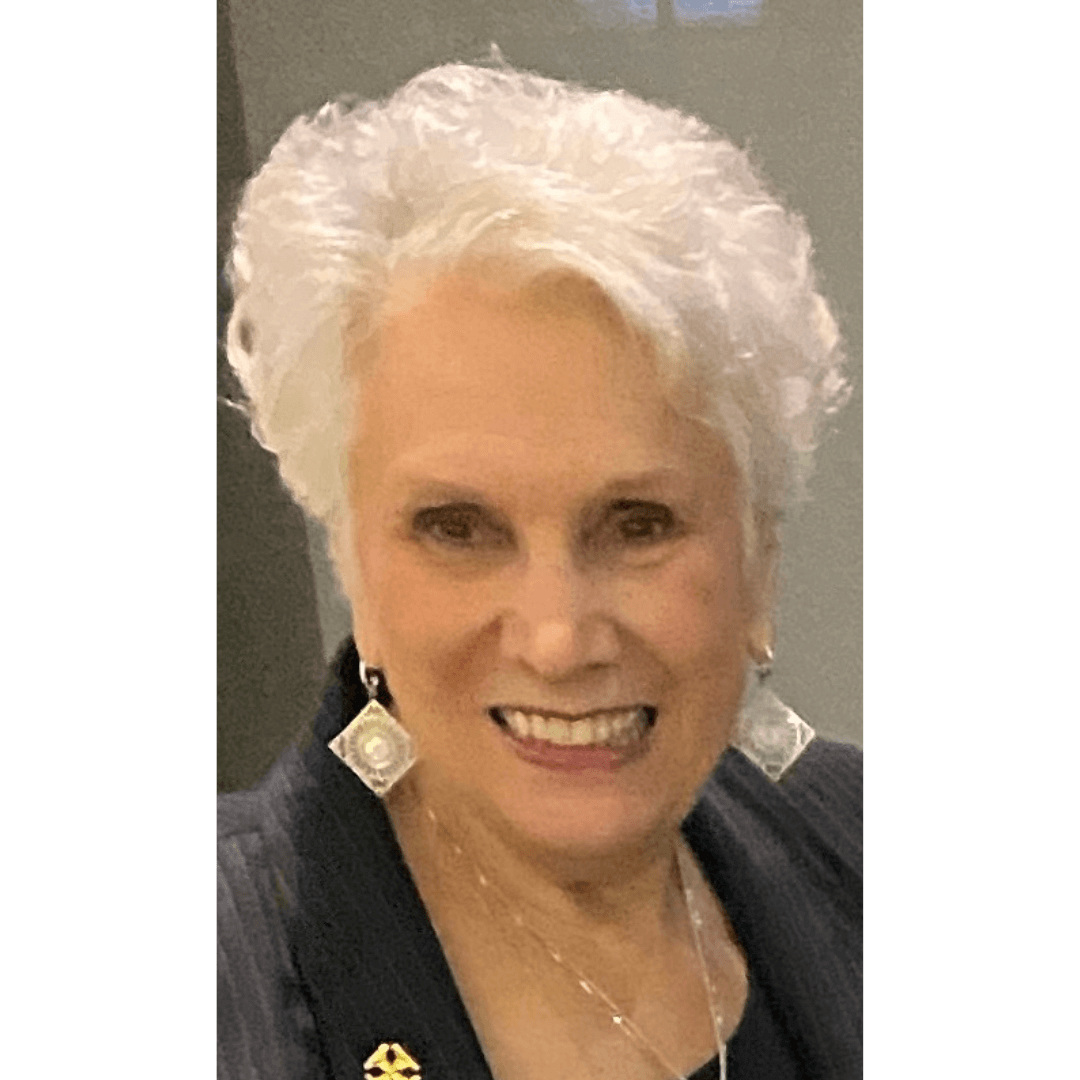 Elderly woman with short white hair smiling, wearing diamond-shaped earrings and a dark blazer.