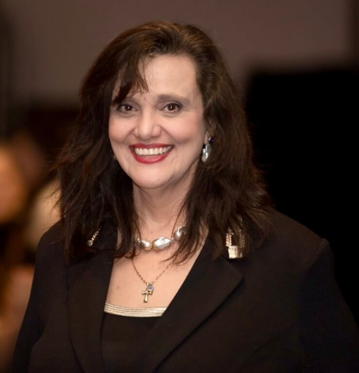 Smiling woman in formal black attire with jewelry, dark hair, red lipstick, and blurred background.