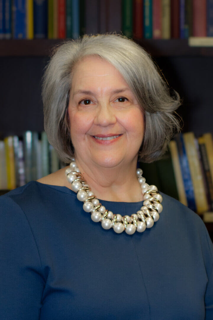 Elderly woman with gray hair, wearing pearl necklace, smiling in front of bookshelf background.