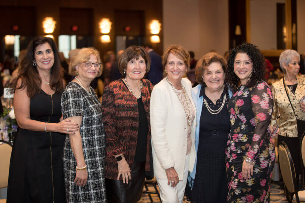 Group of women smiling at a formal event, wearing elegant dresses and standing in a banquet hall.