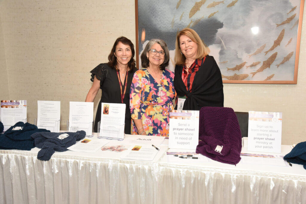 Women at prayer shawl ministry booth with knitted shawls, information pamphlets, and signup sheets at a church event.